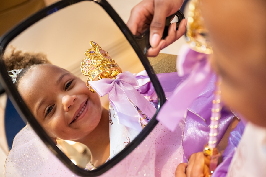 Little girl in a purple Rapunzel princess gown smiling as she sees her crown and wand in a hand mirror at Bibbidi Bobbidi Boutique